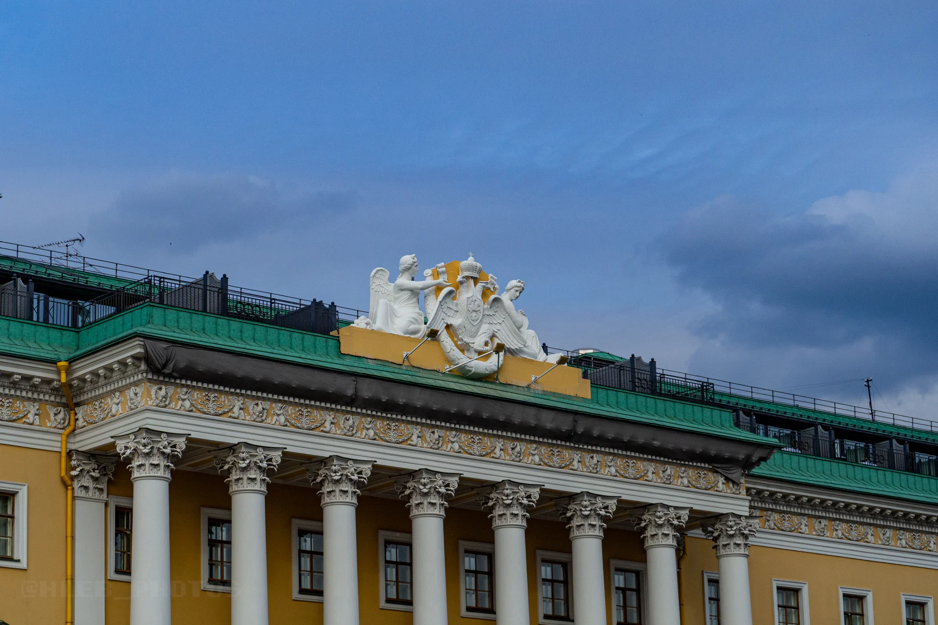 Верхняя часть желтого классицистического здания с рядом белых колонн и скульптурным барельефом на фронтоне.