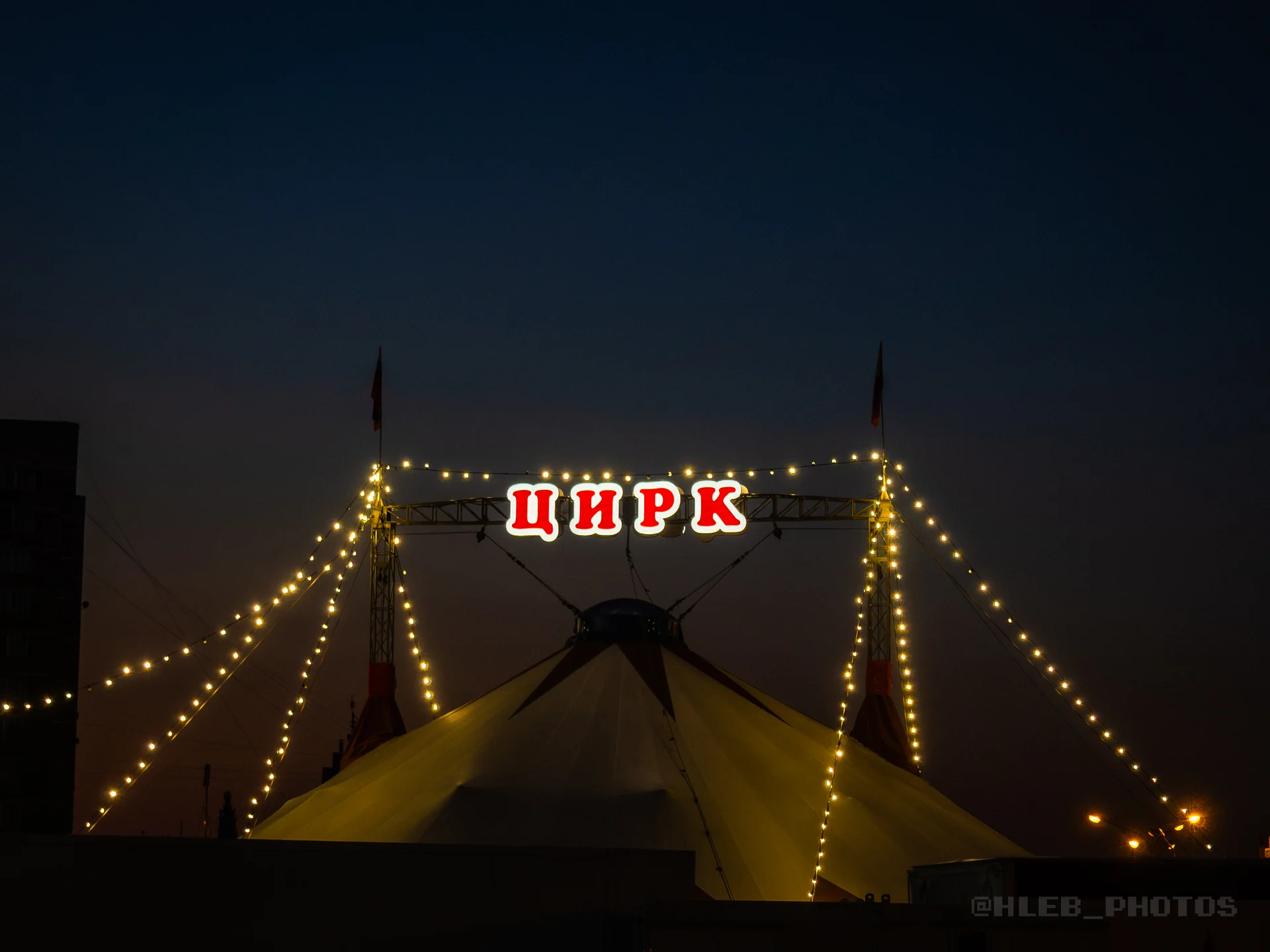Подсвеченный цирковой шатер с надписью «ЦИРК» на фоне темного неба.