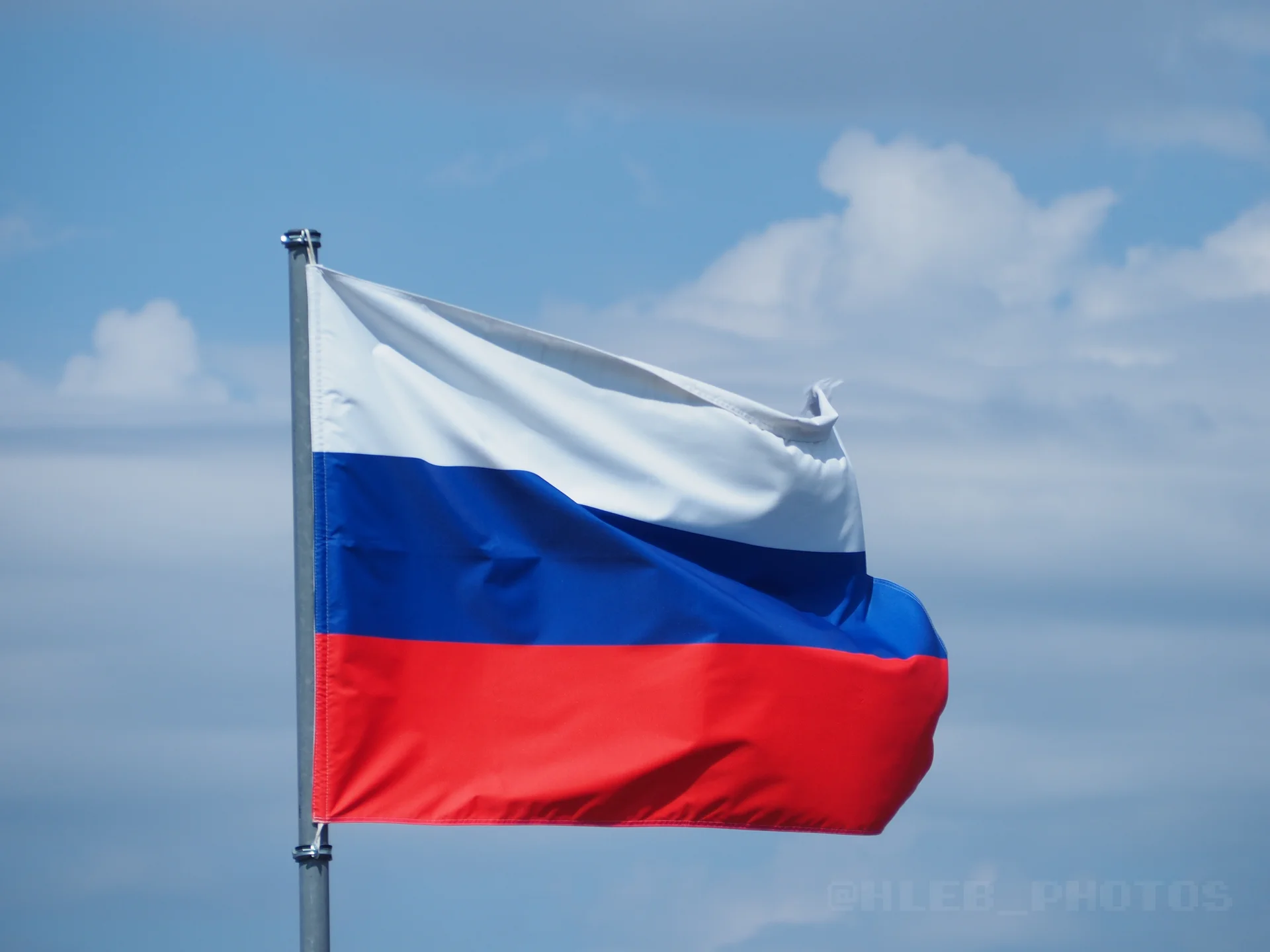 Государственный флаг России развевается на флагштоке на фоне светлого облачного неба.