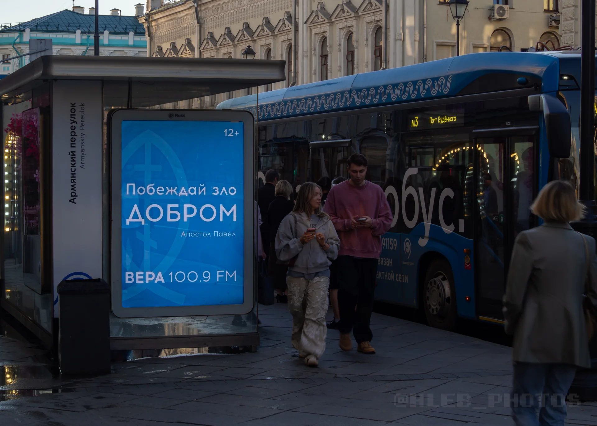 Автобусная остановка со светящимся плакатом "Вера" «Побеждай зло добром» и подъезжающий синий автобус.