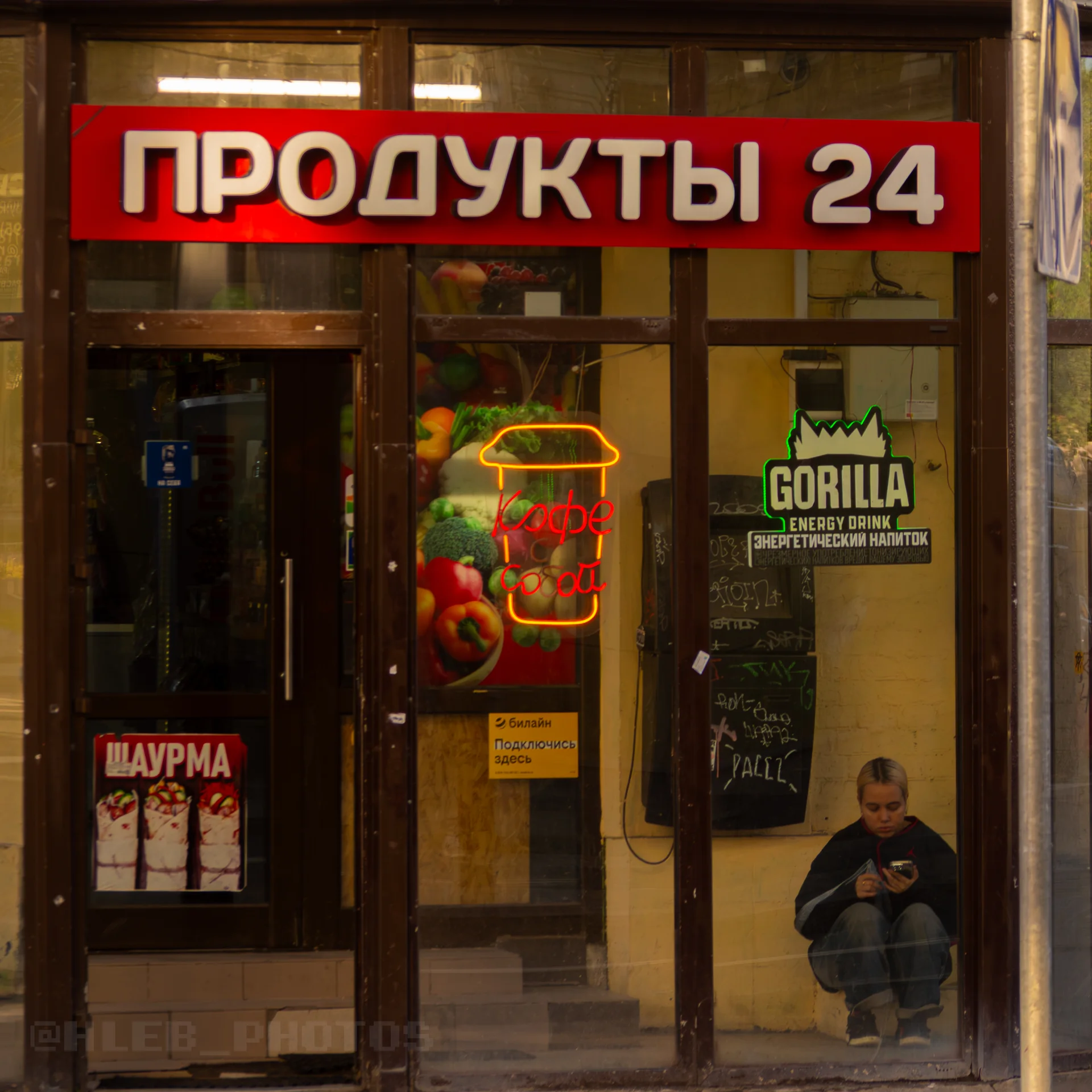 Освещенный вход в круглосуточный магазин «Продукты 24», у витрины сидит человек с телефоном.