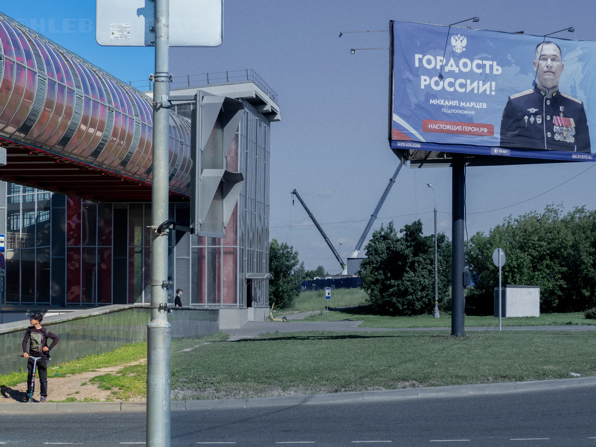 Городской пейзаж с пешеходным мостом, ребенком на самокате и билбордом «Гордость России!» с портретом офицера.