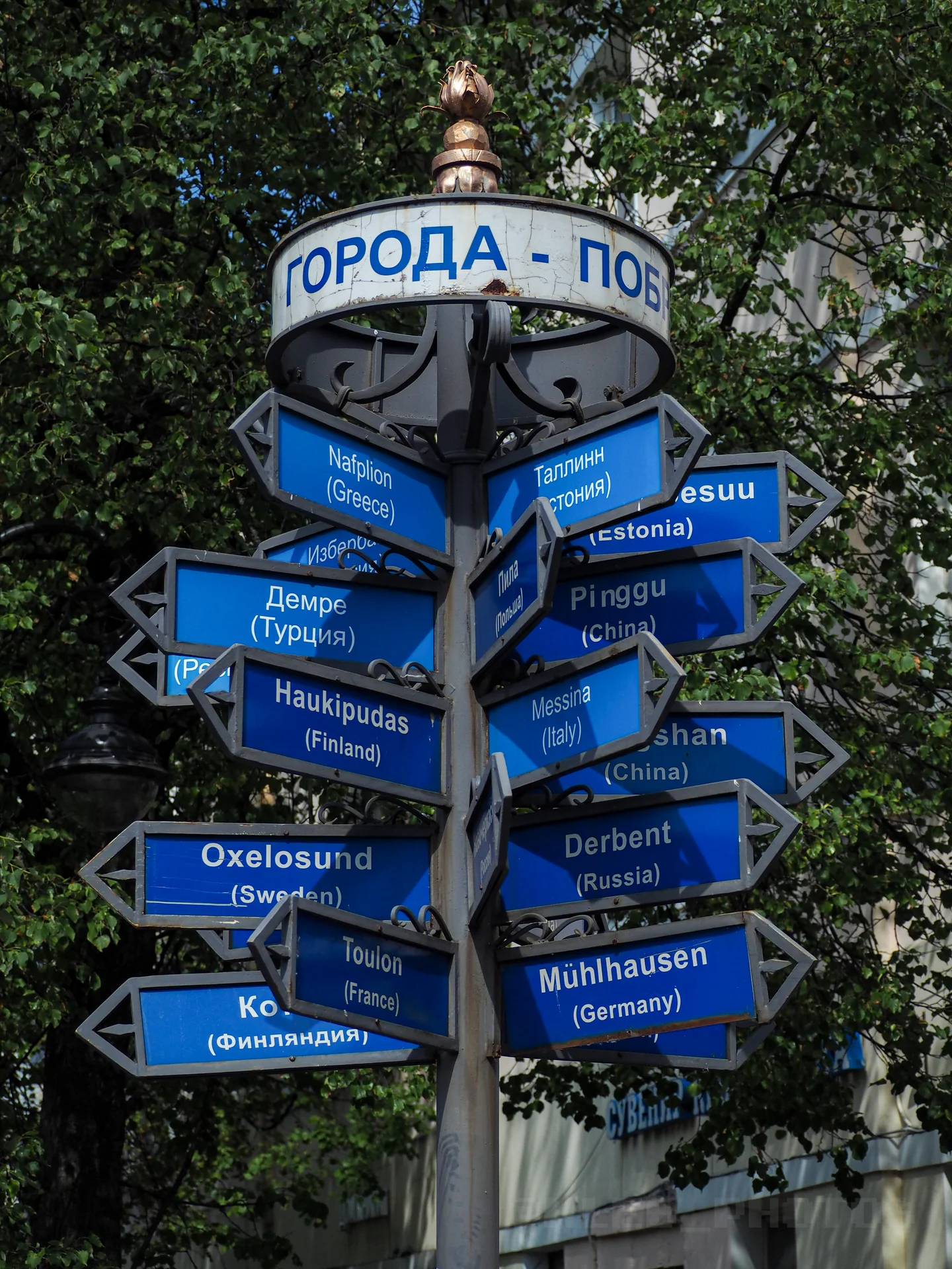 Синий вертикальный указатель с табличками и названиями городов на разных языках на фоне зелени и неба.