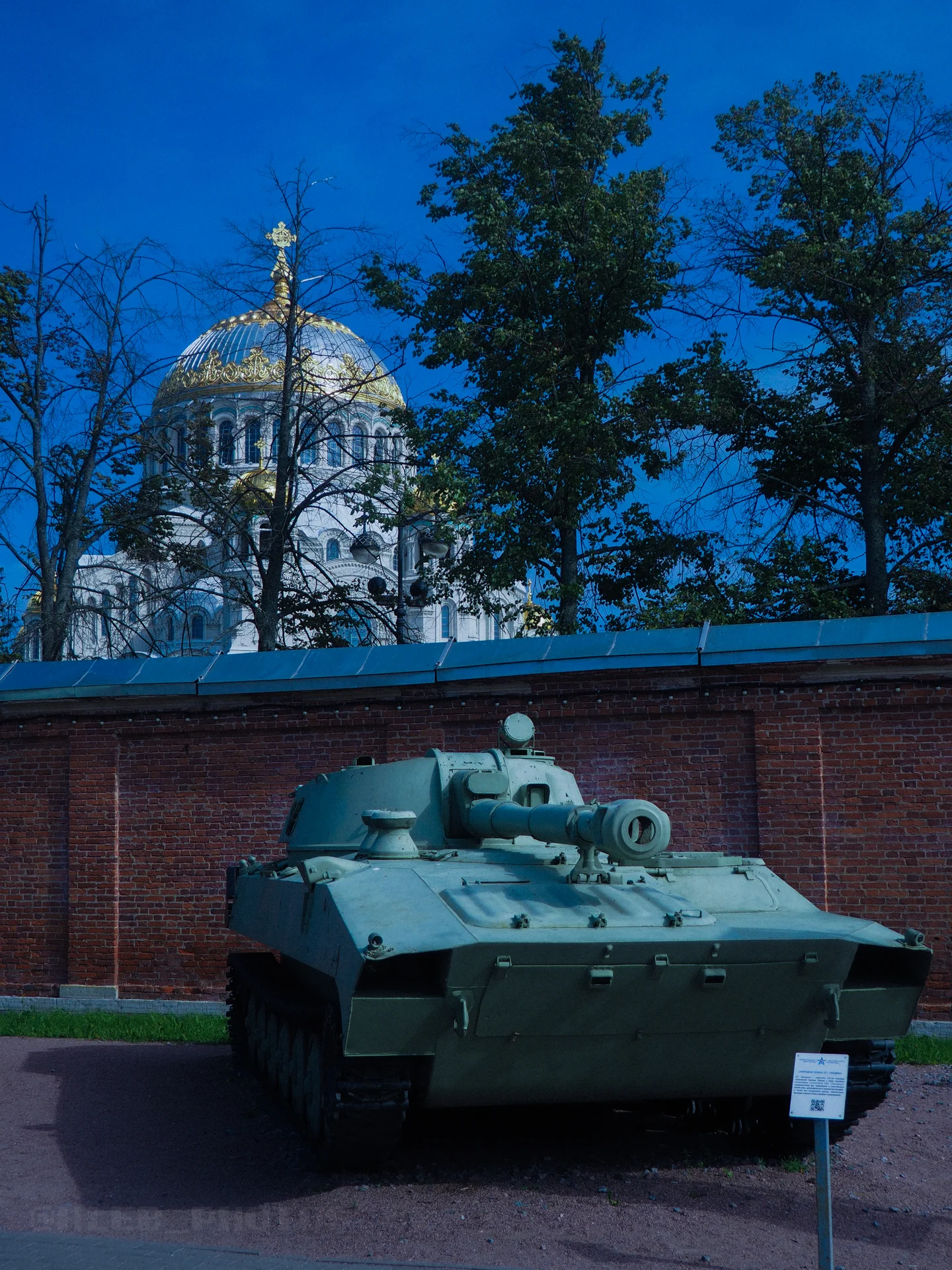 Экспонат военной техники на фоне белого соборного купола с крестом и зеленых деревьев.