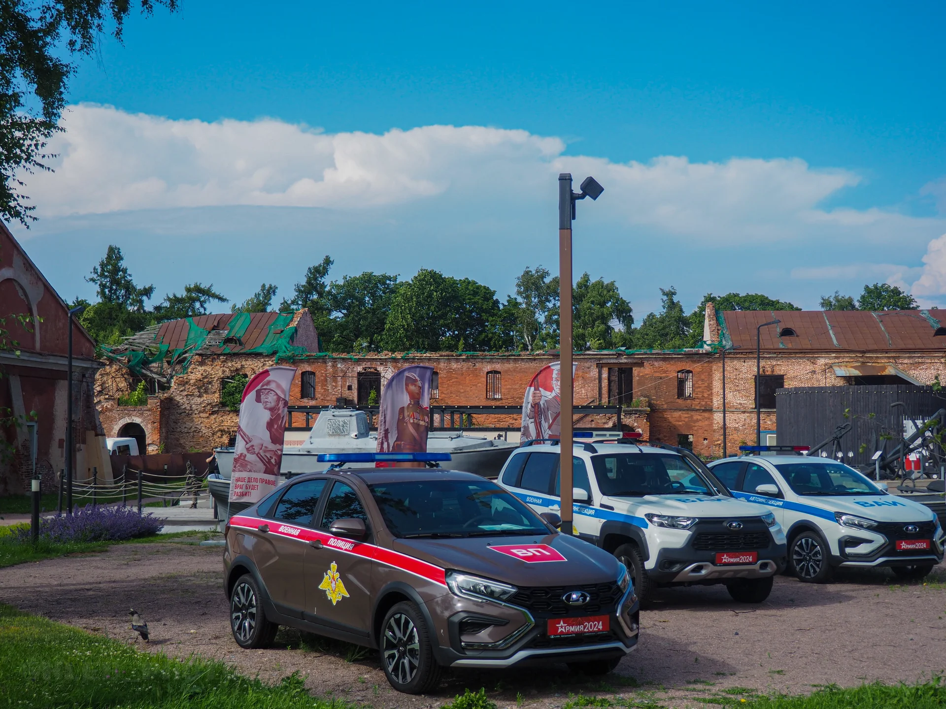 Припаркованные брендированные автомобили спецслужб и полиции на фоне старинного кирпичного здания и деревьев.
