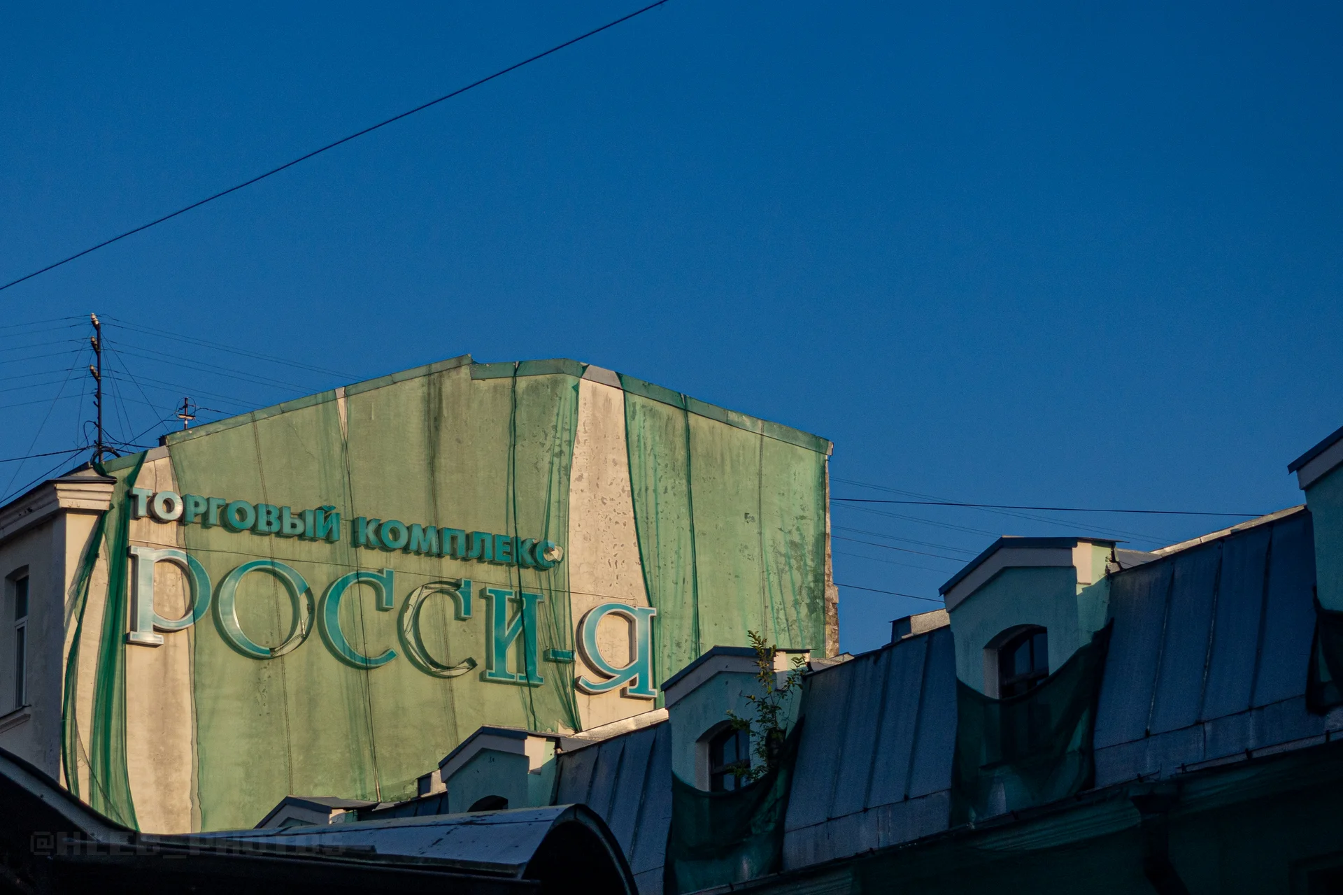 Верхняя часть зелёного здания с крупной вывеской «Торговый комплекс Россия» на фоне синего неба и проводов.