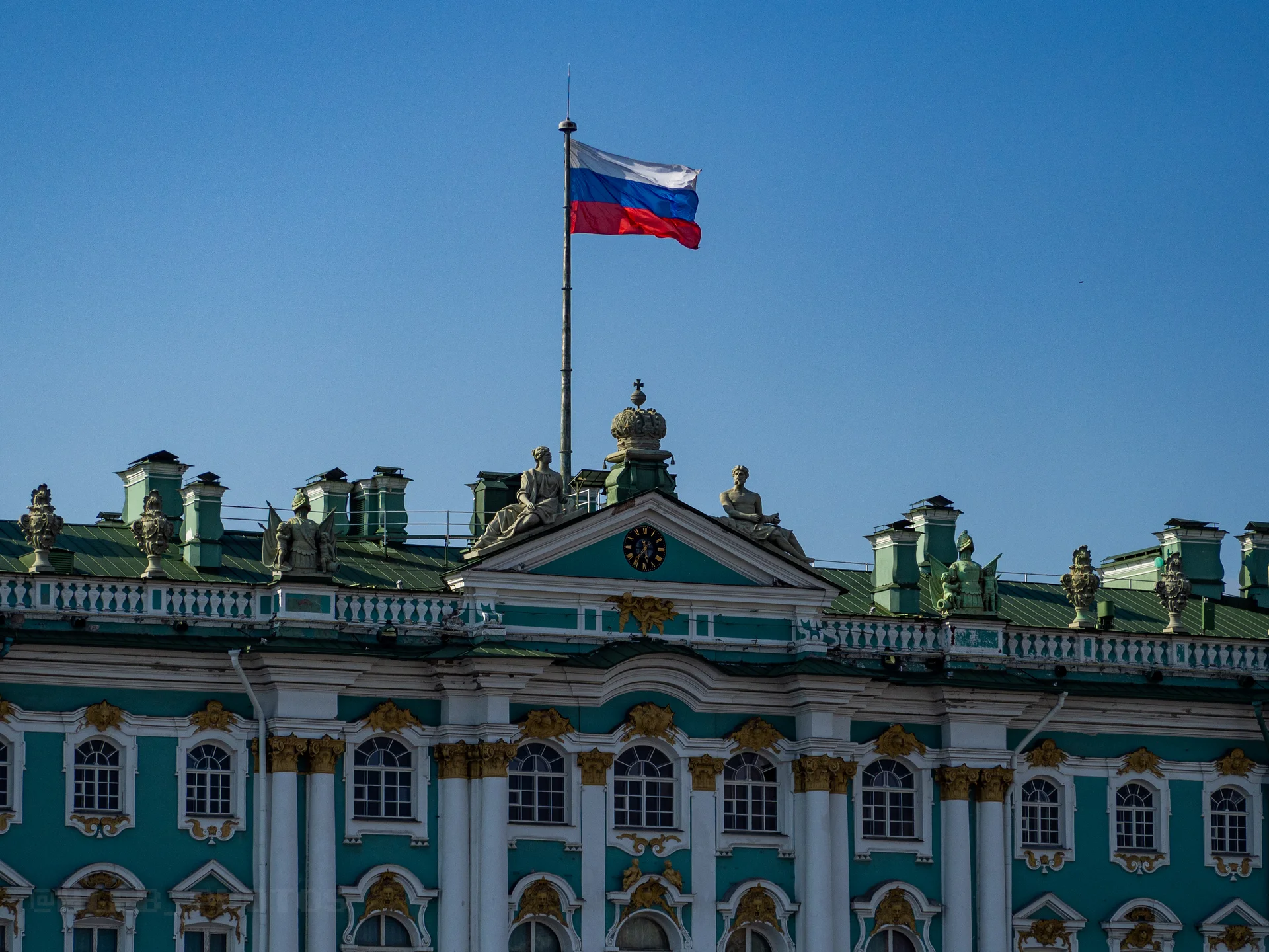 Фасад исторического зеленого здания с белым декором и развевающимся российским флагом на крыше под ярким солнцем.(Зимний дворец СПБ)