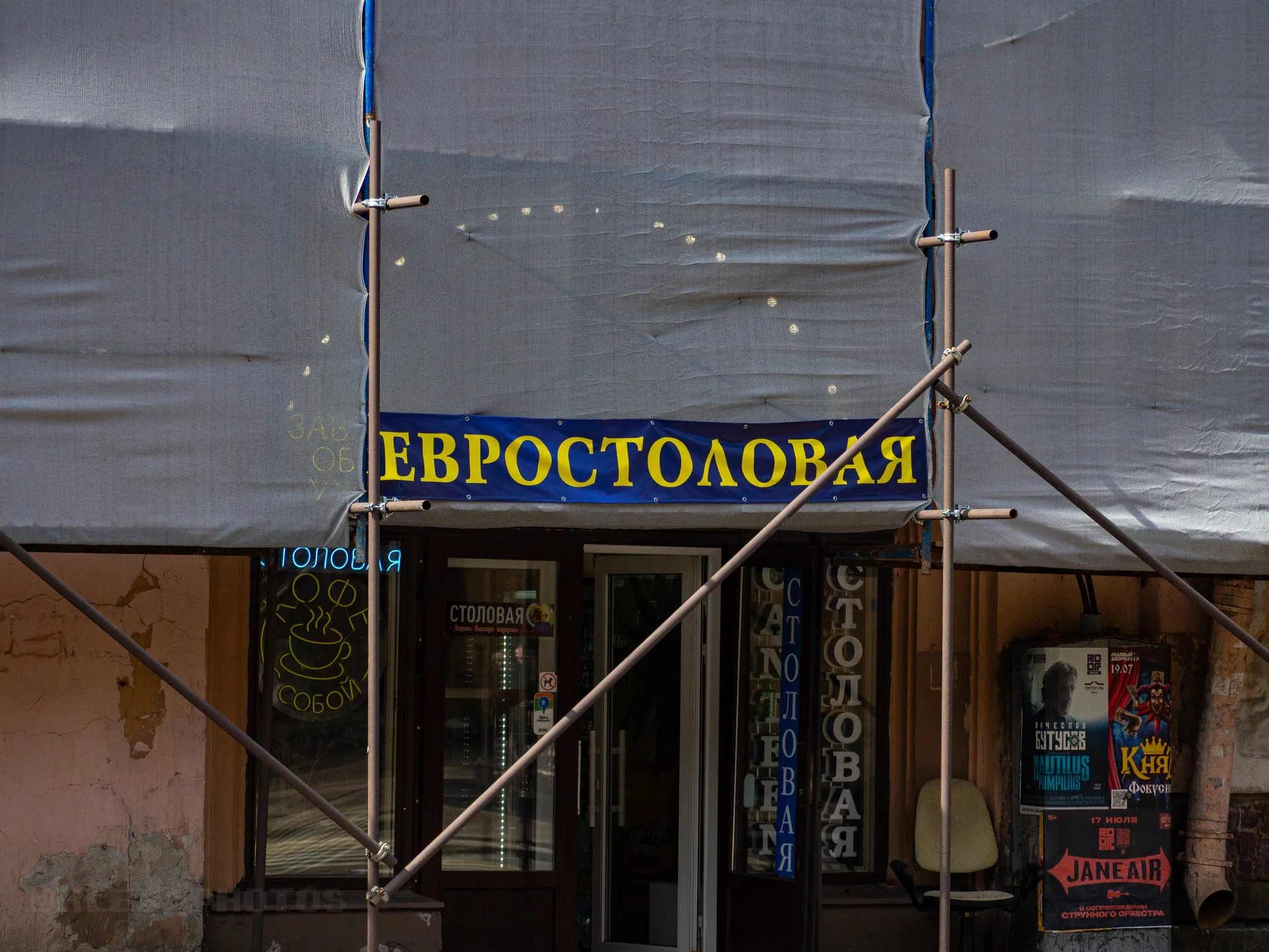 Вход в заведение с желтой вывеской «Евростоловая», закрытый строительными лесами и защитной сеткой на фасаде.