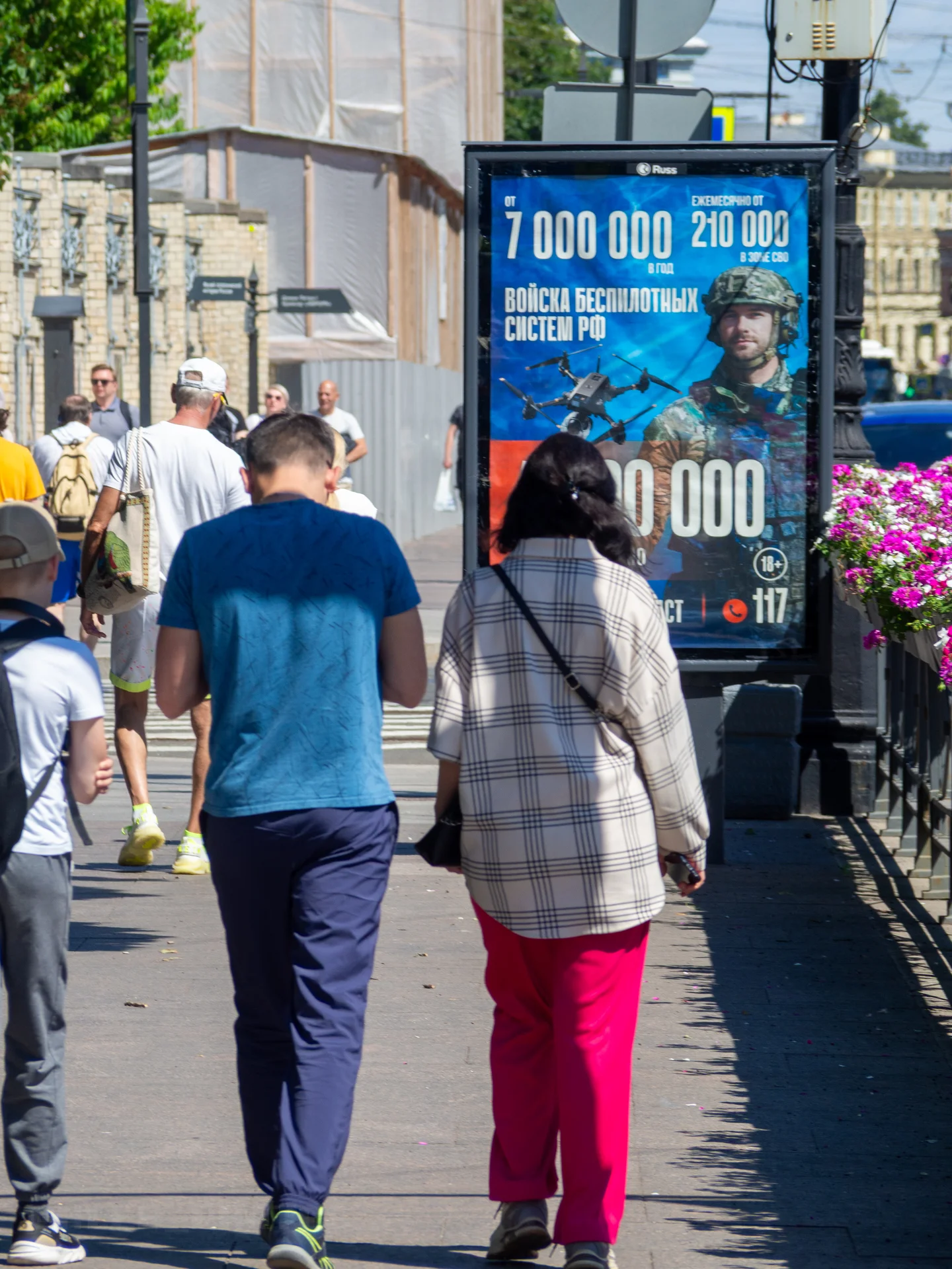 Люди, идущие по улице мимо яркого синего рекламного баннера с изображением военного характера.