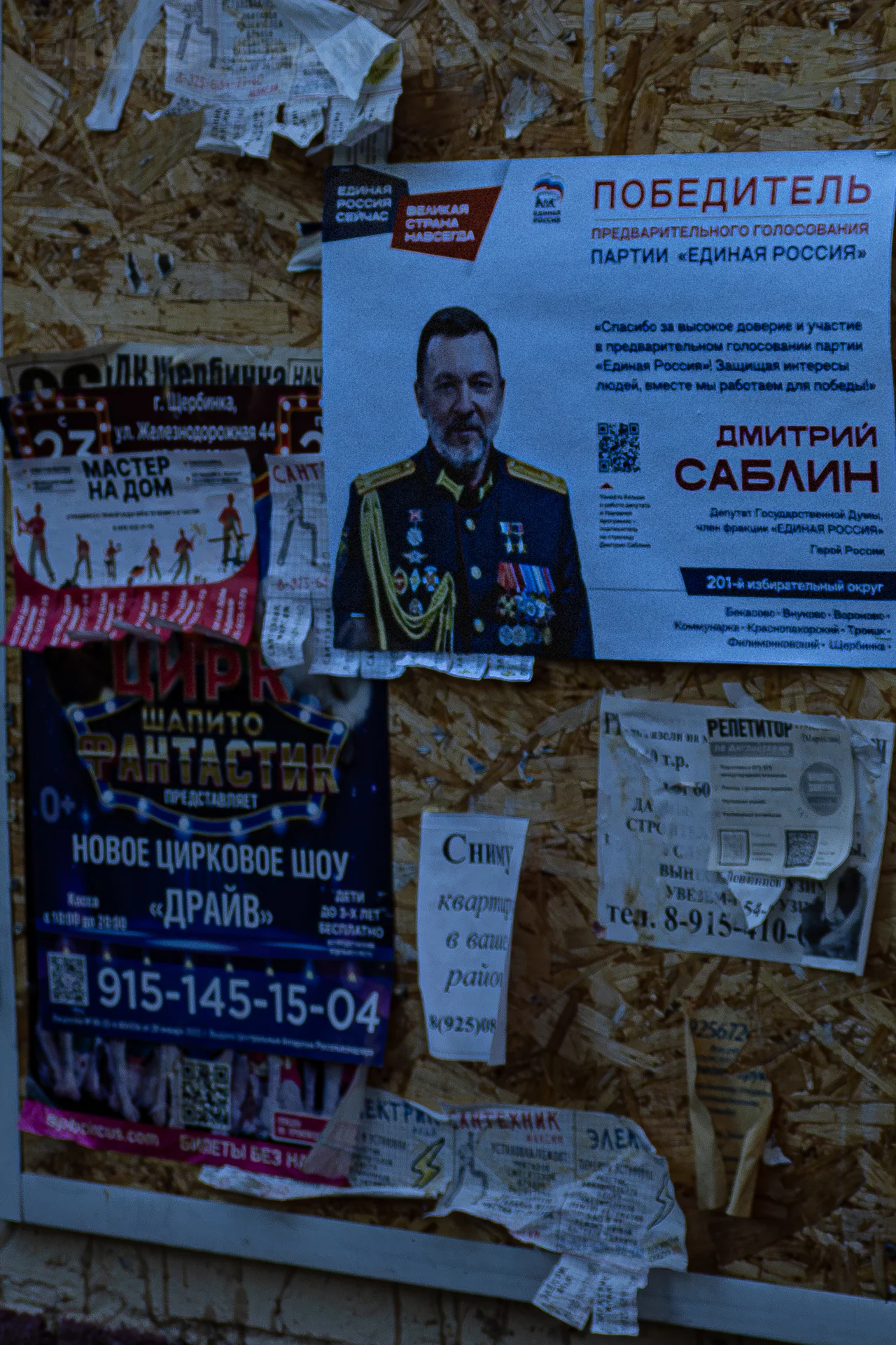 Информационный стенд, плотно оклеенный объявлениями, включая политическую листовку Единой России и рекламу циркового шоу.