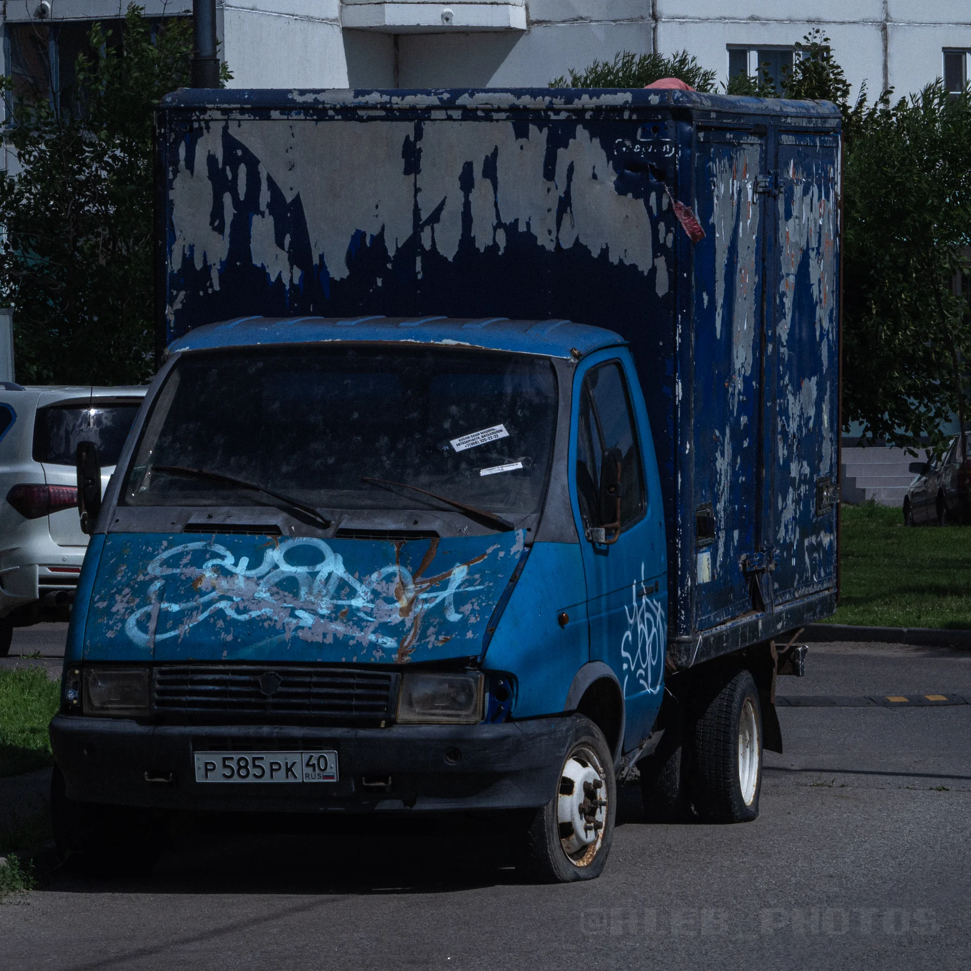 Старый синий грузовой фургон с облупившейся краской и граффити на кабине, припаркованный на улице.