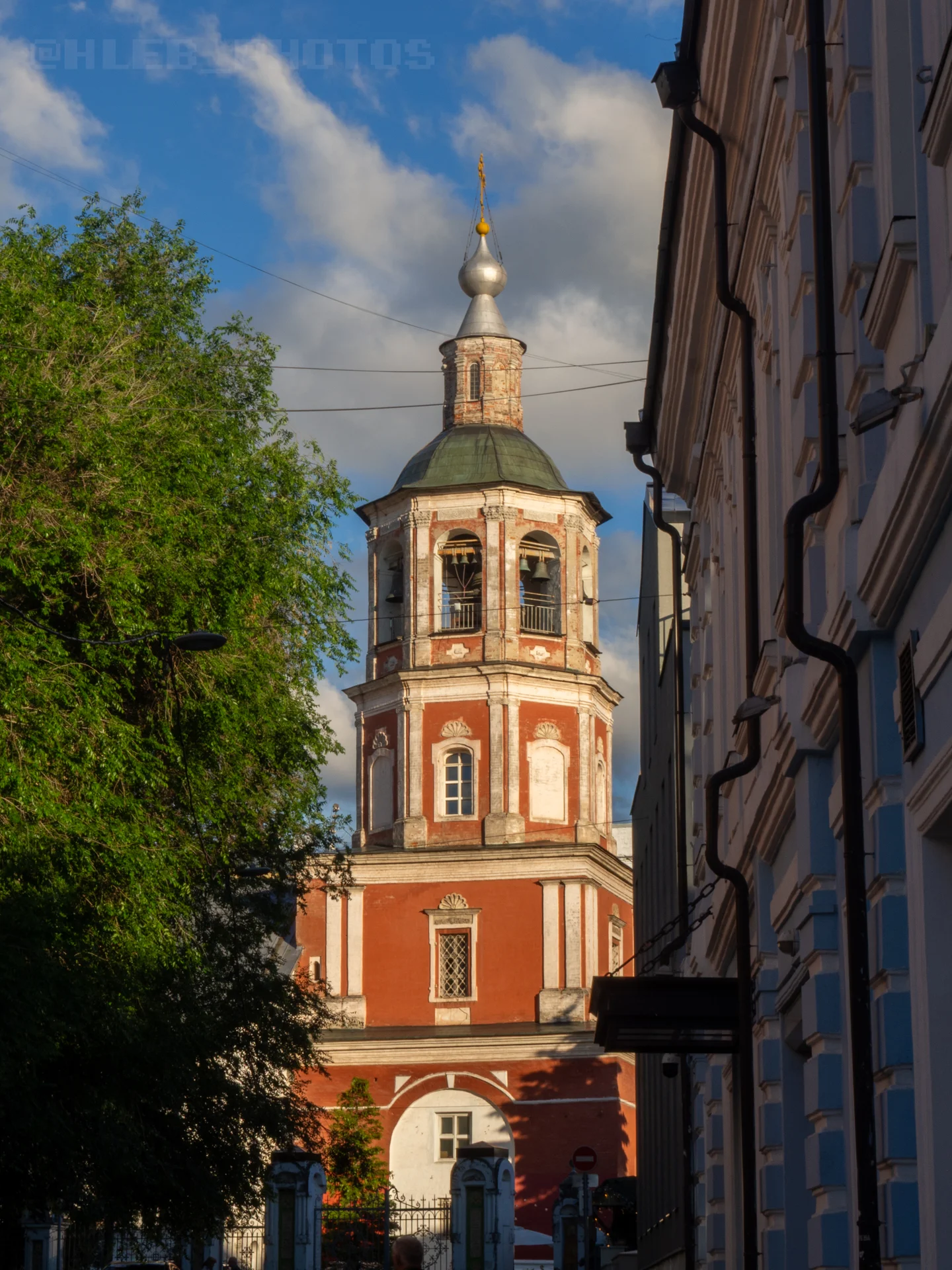 Высокая церковная колокольня из красного кирпича в окружении деревьев и зданий.