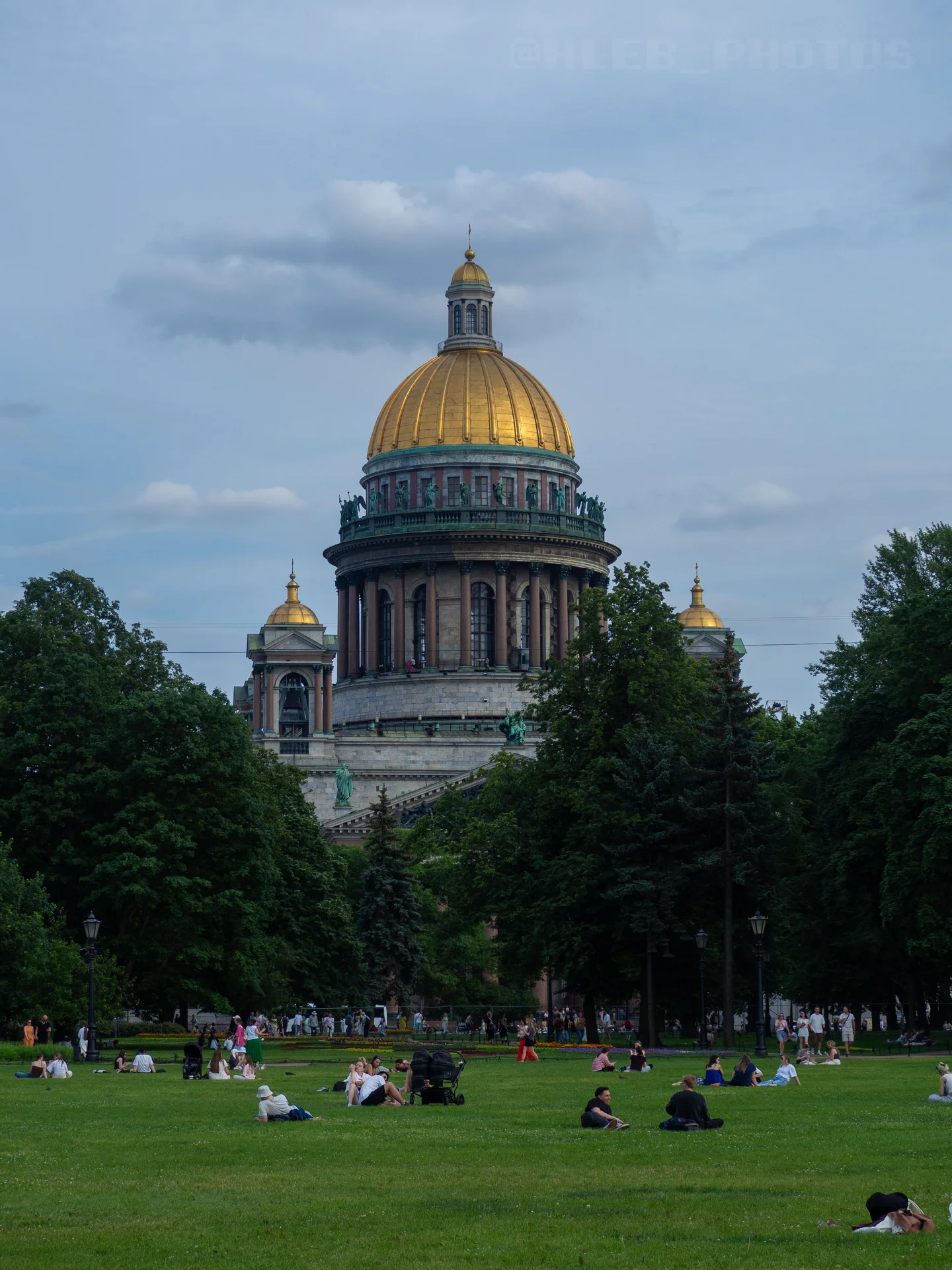 Купол Исаакиевского собора с золотой главой на фоне зеленой листвы деревьев и людей, отдыхающих на газоне.