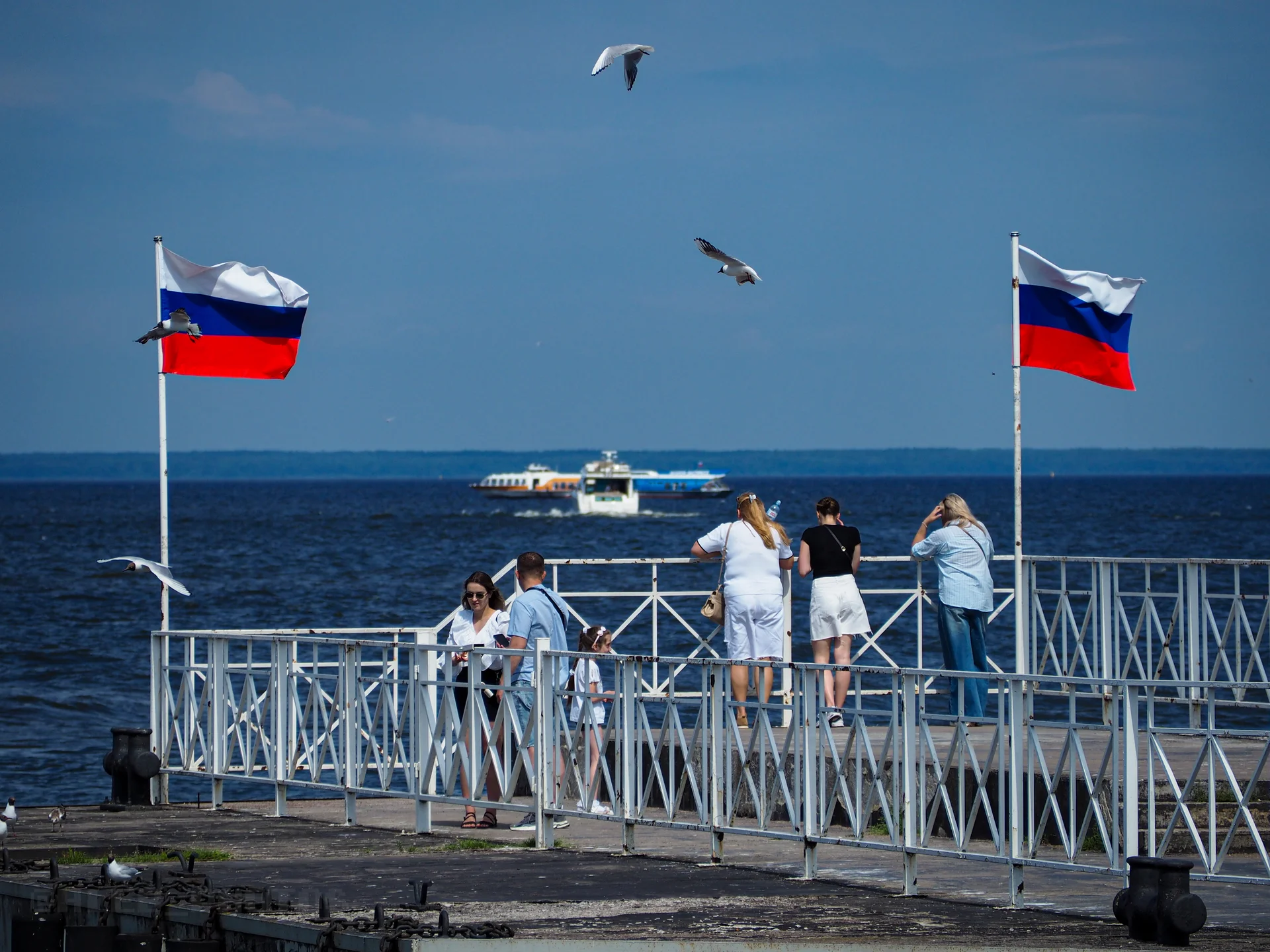 Люди на фоне водной глади, плывущего катера и развевающихся российских флагов на набережной.