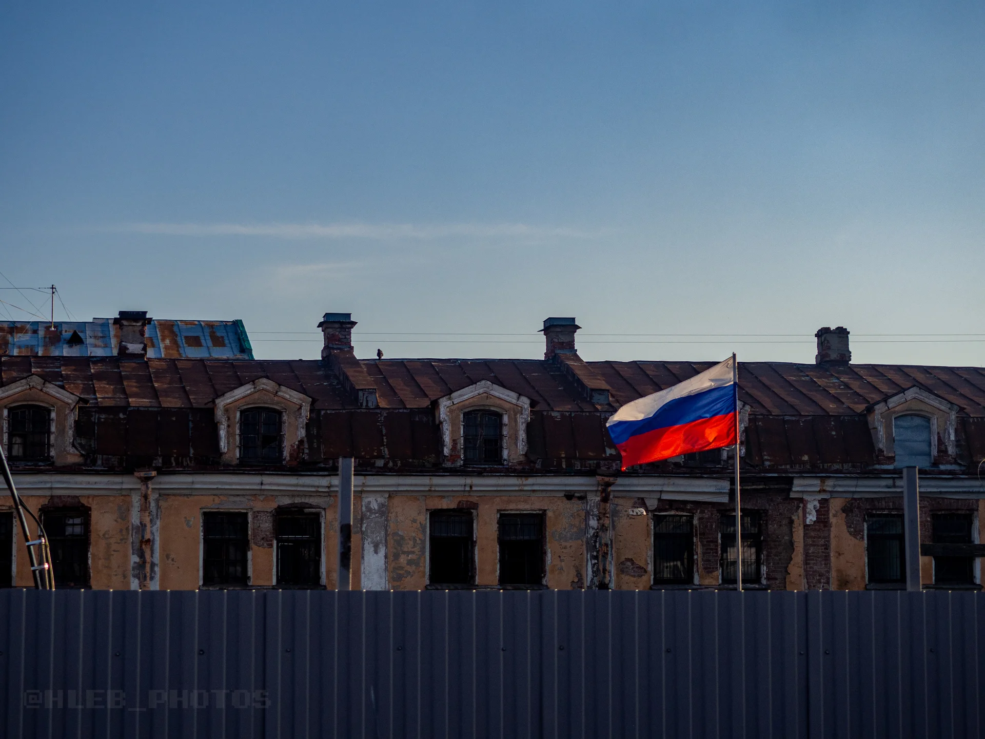 Верхние этажи и крыши старинных зданий за серым забором, над которым развевается российский флаг.
