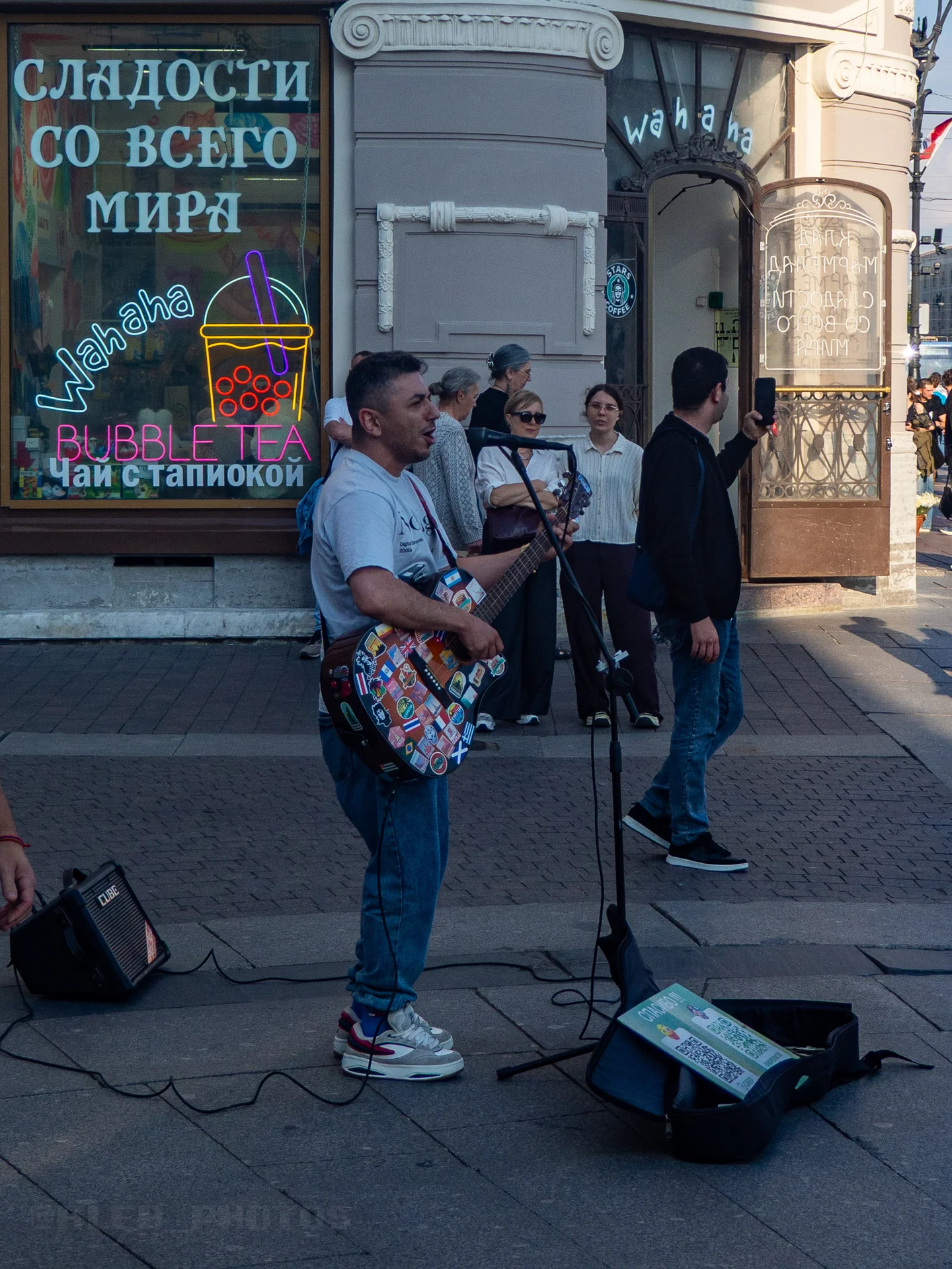 Мужчина с гитарой выступает на улице перед зрителями у здания с витражной витриной.